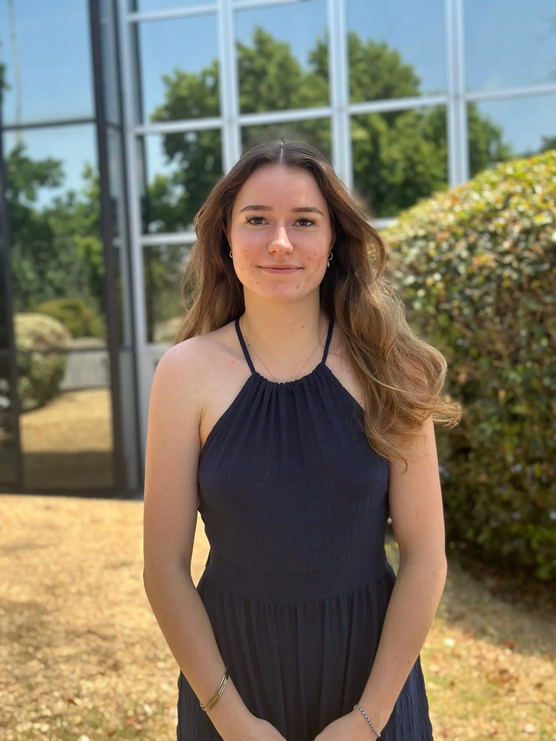 Portrait d'Alyah Mathieu en robe bleu marine devant une haie et une façade vitrée.