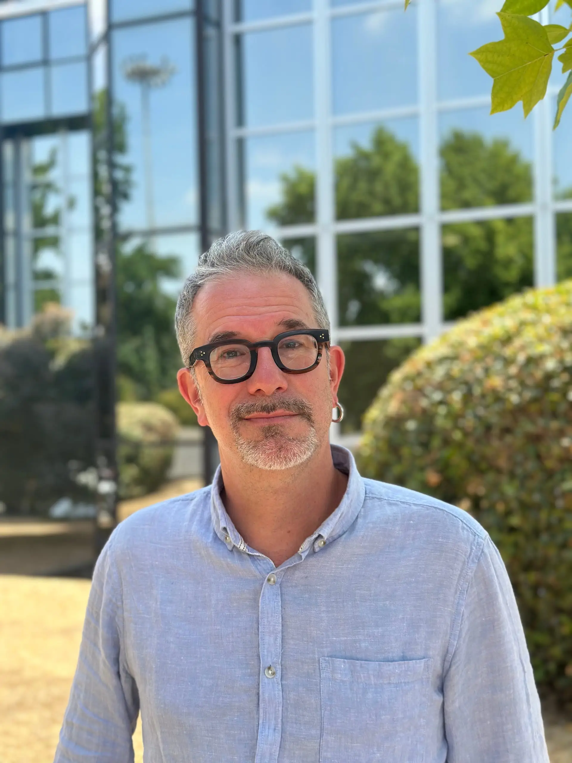 Portrait de Pierre Coustures avec des lunettes noires devant une haie et une façade vitrée.