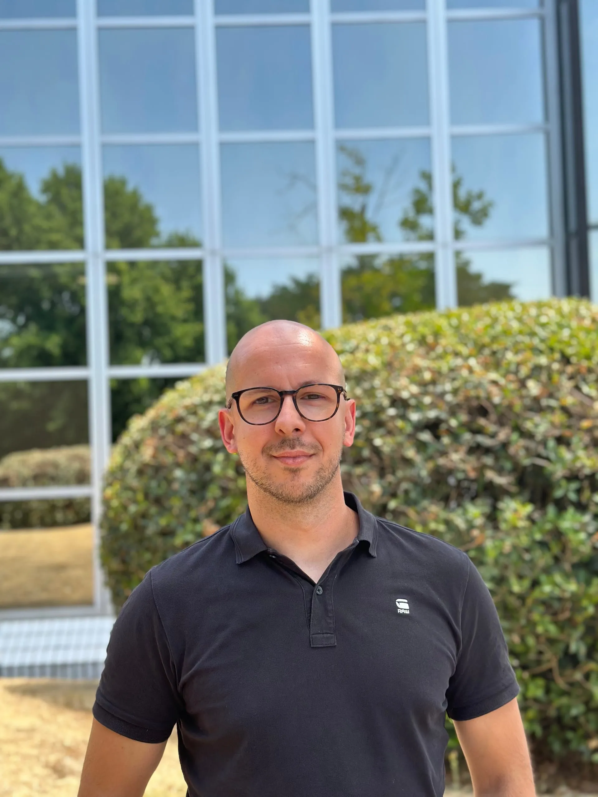 Portrait de Thomas Triolo avec des lunettes noires devant une haie et une façade vitrée.